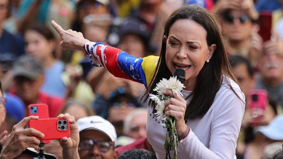 María Corina Machado s'adressant à une foule, tenant des fleurs blanches, symbole de son leadership et de sa lutte pacifique pour la démocratie au Venezuela.