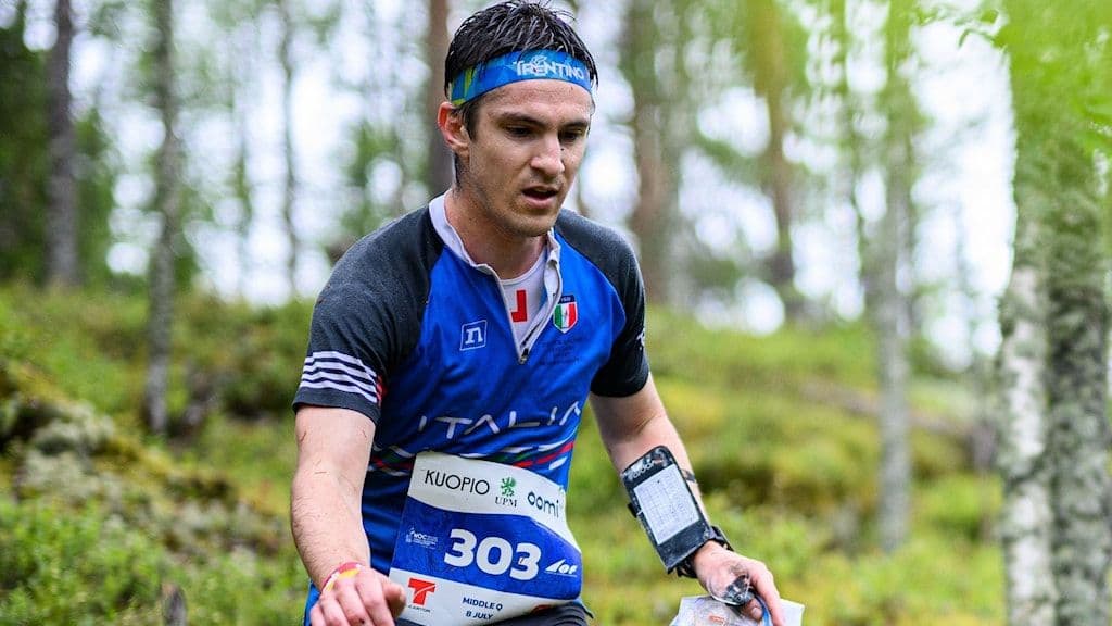 Mattia Debertolis, an Italian orienteer, runs through a forest during a competition, holding a map and wearing bib number 303.