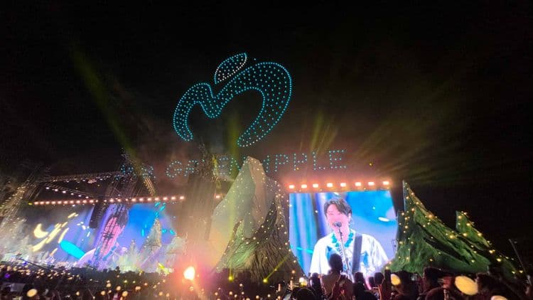 Mrs. GREEN APPLE performing live on a large stage with vibrant light effects and a large screen displaying the vocalist, illustrating their dynamic sound.