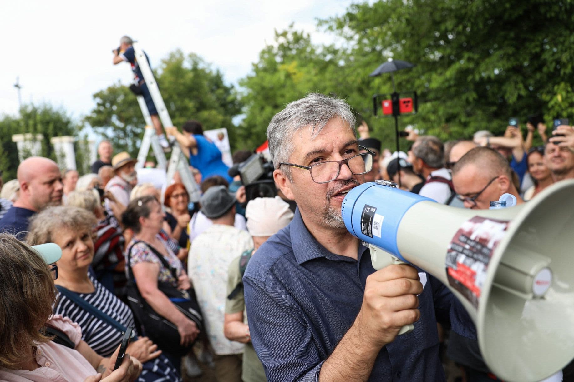 Opposition MP Ákos Hadházy addressing protesters with a megaphone, with people on ladders in the background at the Hatvanpuszta estate.