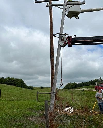 Ouvrier réparant un poteau électrique penché dans un champ, illustrant les dégâts causés par les phénomènes météorologiques extrêmes.