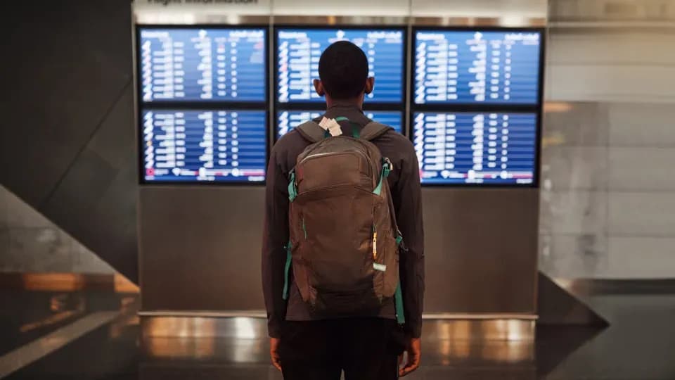 Passageiro olhando um painel de voos em aeroporto, representando os direitos dos viajantes afetados por atrasos e cancelamentos.