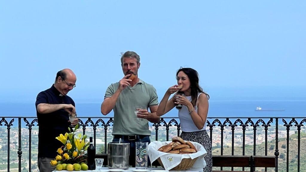 People enjoying a meal outdoors with a scenic view of Milazzo and the sea, showcasing the region's diverse culinary landscape and authentic flavors.