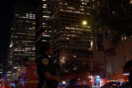Police officer standing in front of illuminated New York City skyscrapers at night, symbolizing the enduring power and security of physical locations in finance