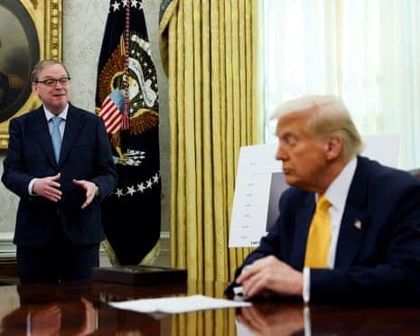 President Donald Trump seated at a desk while an economic official stands, discussing economic data in an office.