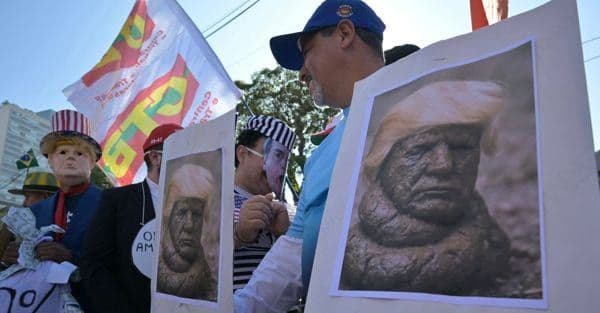 Protesters holding signs depicting Donald Trump's face as a weathered bust, symbolizing the impact of his trade policies.