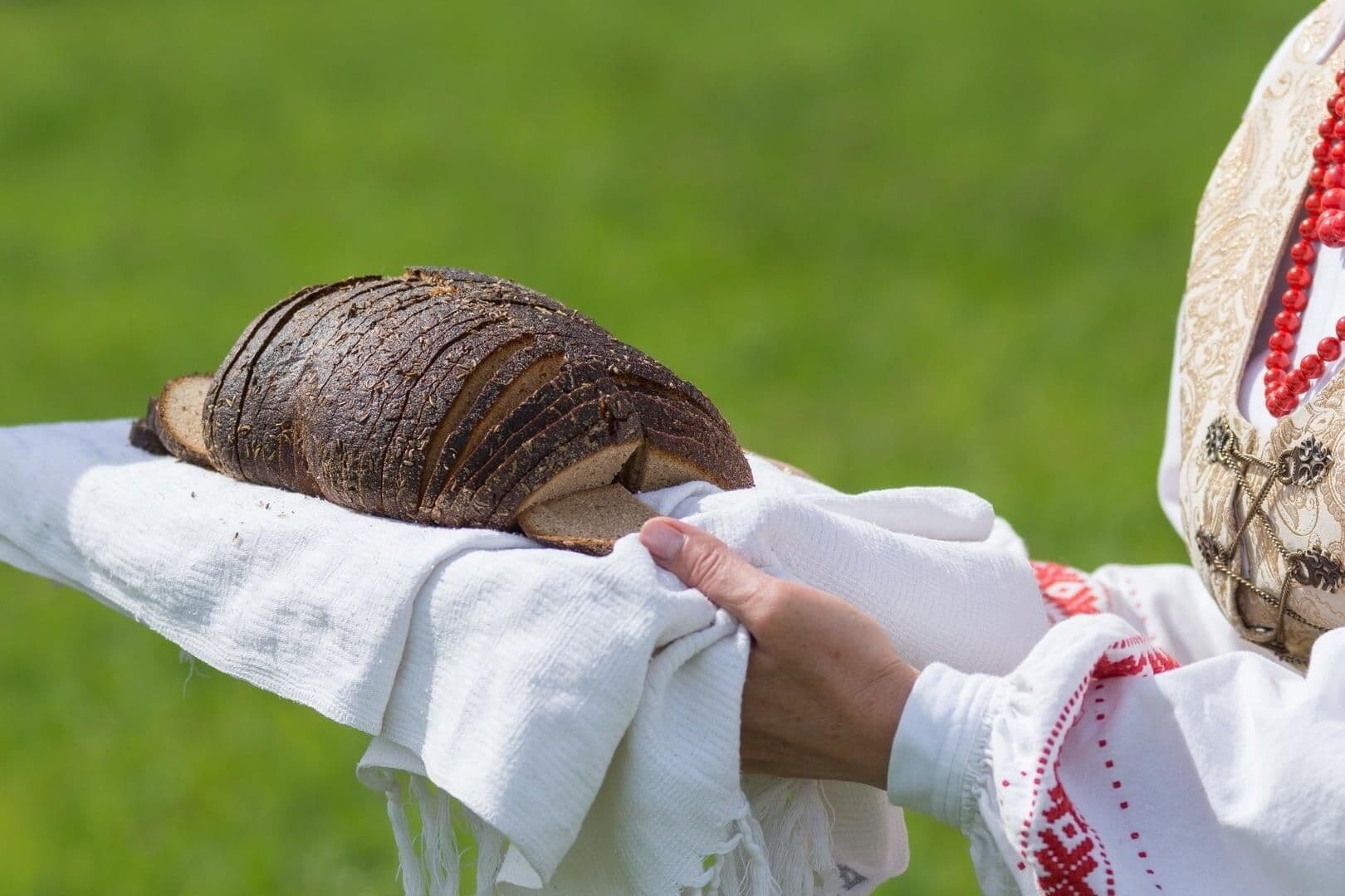 Rankos, apsirengusios tradiciniais drabužiais, laiko supjaustytą ruginės duonos kepalą, pabrėžiantį jos svarbą kasdienėje mityboje.