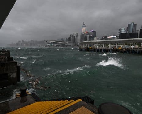Rough waves crash against a pier in Hong Kong's Victoria Harbour under dark, stormy skies, with the city skyline in the background, illustrating the impact of a severe typhoon.
