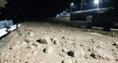 Route recouverte de boue et de roches après une coulée de boue nocturne, avec un pont en arrière-plan, illustrant les dégâts sur les infrastructures.