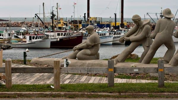 Statues of labourers pulling ropes by a harbour with fishing boats, symbolizing the collective effort and struggle of migrant workers towards genuine empowerment.