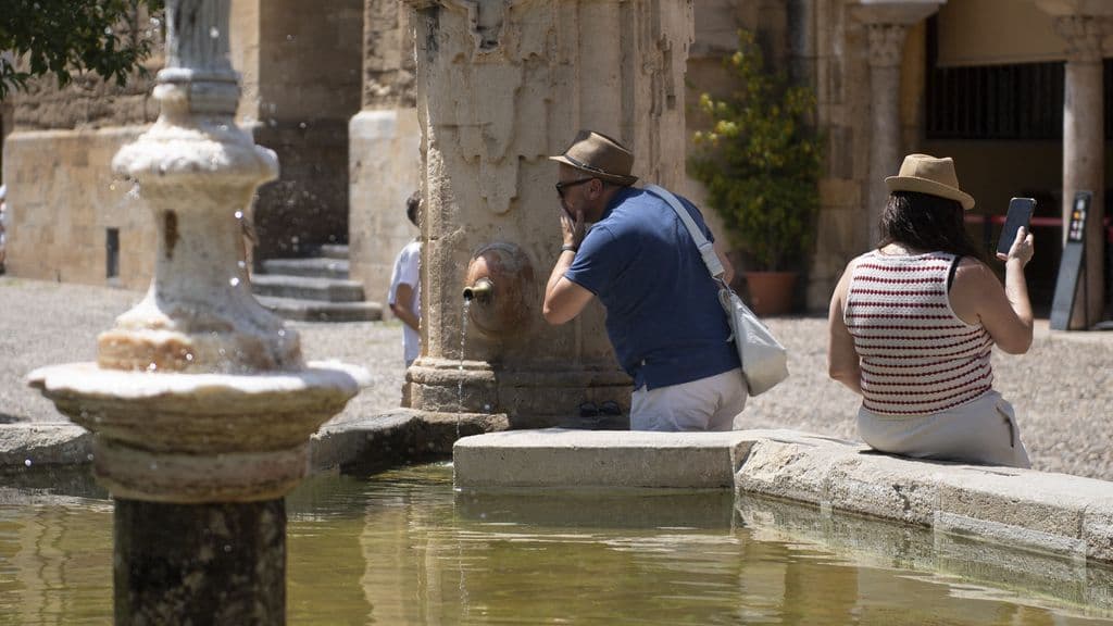 Tourists seeking relief from the scorching sun by a fountain in a historic Spanish city, one drinking water and another resting.