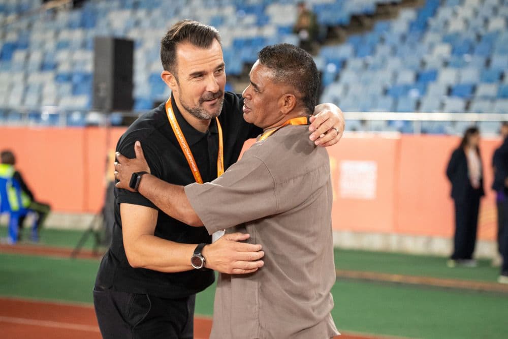 Two football coaches or officials embracing on the sidelines of a stadium, symbolizing collaboration and camaraderie at CHAN 2025.