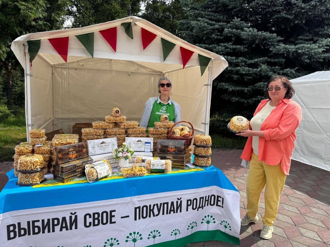 Two women at an outdoor market stall selling traditional pastries, with a banner encouraging local purchases, symbolizing community trade.