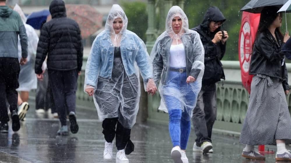 Two women in clear plastic ponchos walking across a bridge in London during heavy rain, illustrating urban impact of severe weather.