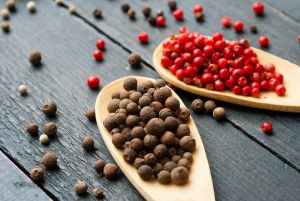 Two wooden spoons holding different types of peppercorns, one with black and another with red, on a dark wooden surface.