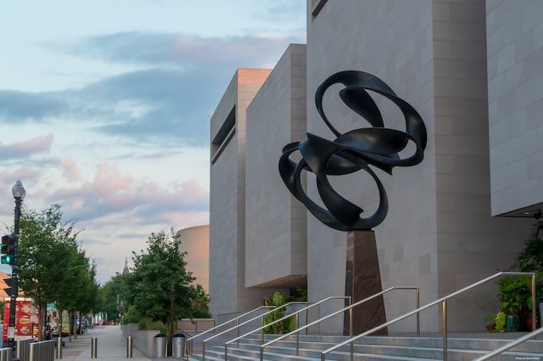 Un bâtiment moderne sous un ciel nuageux, avec une sculpture abstraite au premier plan, représentant le ciel urbain et ses variations.
