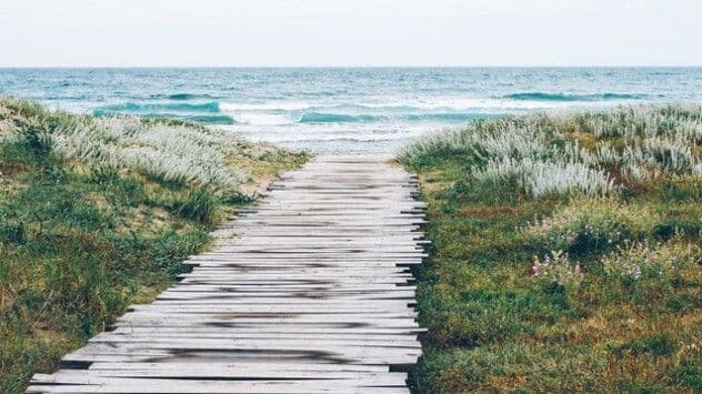 Un chemin en bois menant à une plage et à la mer sous un ciel nuageux, symbolisant l'adaptation des loisirs aux caprices de la météo estivale.