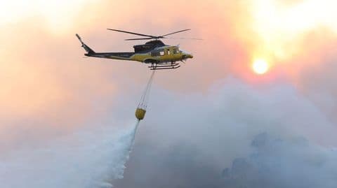 Un hélicoptère bombardier d'eau largue de l'eau sur un incendie de forêt à Ponteceso, avec le soleil couchant en arrière-plan.