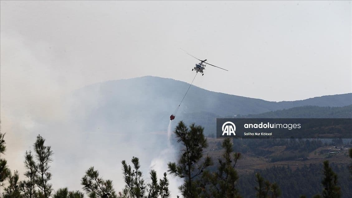 Un hélicoptère de lutte contre les incendies largue de l'eau sur un feu de forêt fumant dans une zone montagneuse.