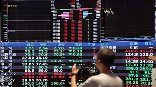 Un homme filmant un écran d'affichage boursier, symbolisant l'observation des tendances du marché et les perspectives d'avenir pour l'industrie des semi-conducteurs.