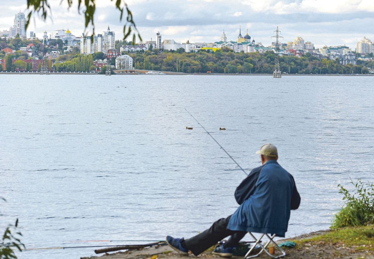 Un homme pêchant tranquillement au bord d'une rivière à Voronezh, avec la ville en arrière-plan, symbolisant l'impact de la météo sur la vie quotidienne.