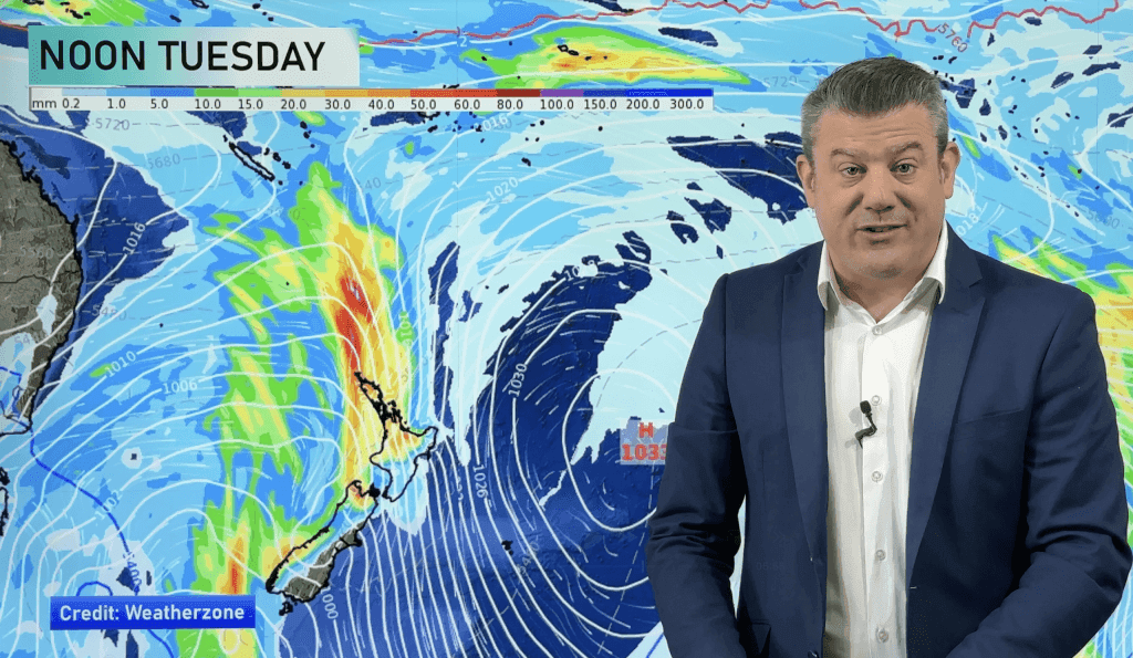 Un présentateur météo devant une carte de Nouvelle-Zélande montrant un système de pluie intense prévu pour mardi.