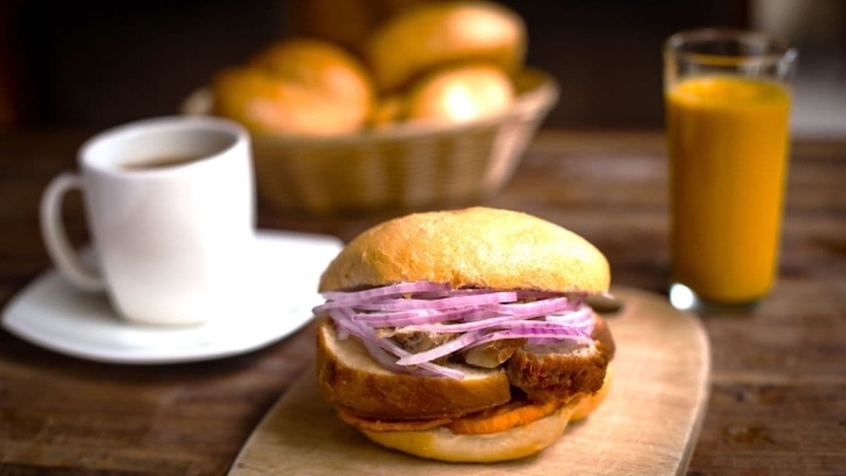 Un tradicional pan con chicharrón peruano con cebolla, acompañado de café y jugo en una mesa de desayuno