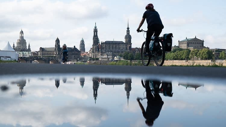 Une personne à vélo sur une piste cyclable, avec le reflet d'une ville historique et du ciel dans une grande flaque d'eau, symbolisant l'été incertain entre soleil et orages.