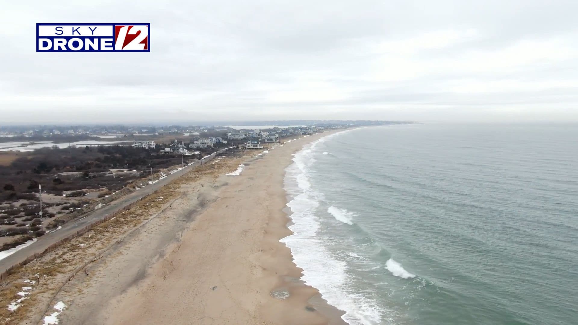 Vue aérienne d'une longue plage de sable et de l'océan sous un ciel couvert, soulignant la beauté naturelle du temps.
