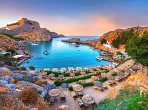Vue panoramique d'une baie grecque pittoresque avec plage, bateaux et bâtiments historiques au coucher du soleil, symbolisant les merveilles de la Grèce.